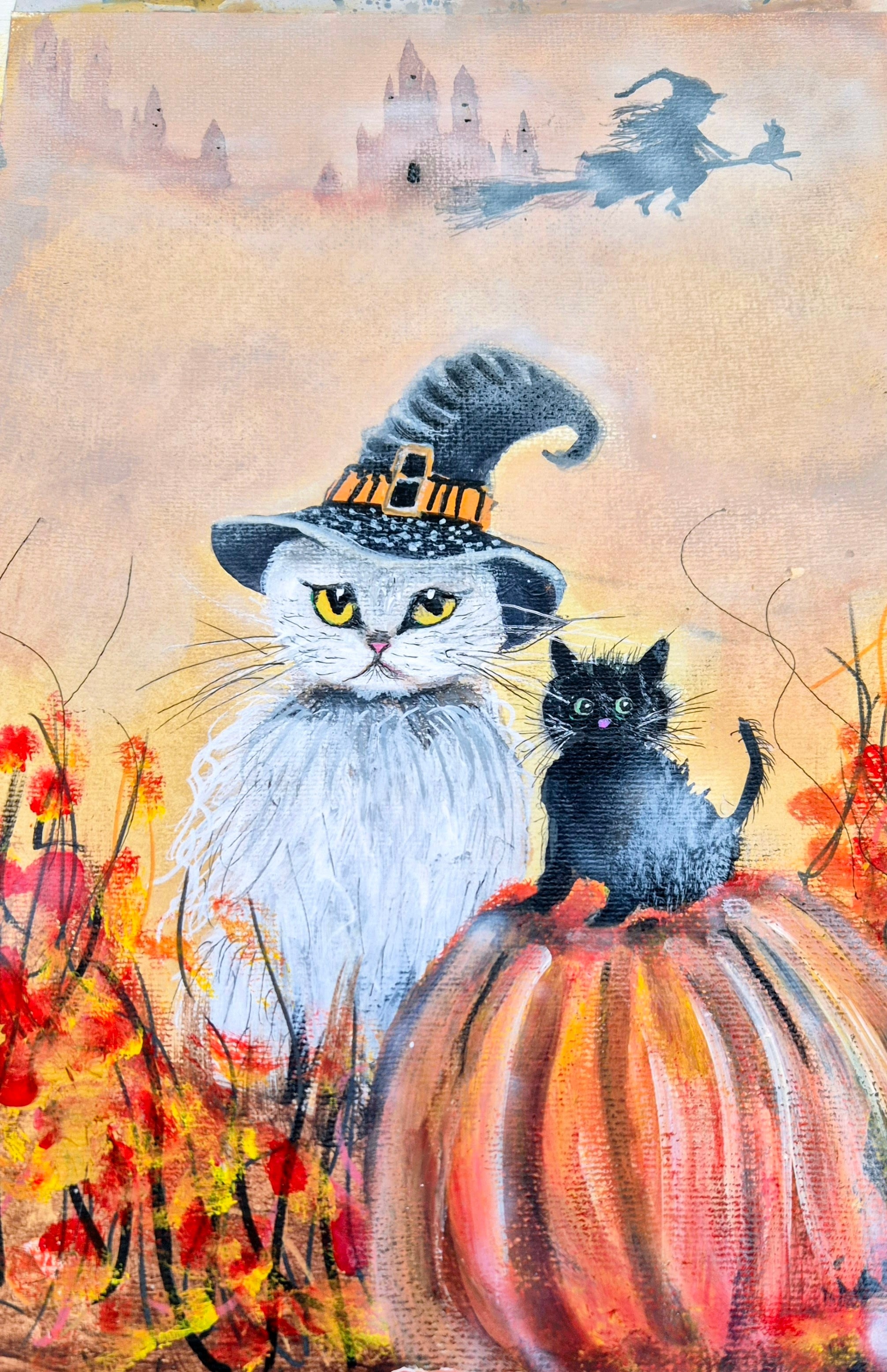 White cat wearing a speckled witch’s hat with golden buckle beside a curious little black kitten perched on a great pumpkin — warm autumn fields with fiery red and orange foliage, misty distant castle towers and a witch flying past on her broomstick across soft golden skies — original whimsical Halloween art by Janet Jackson, JJs Art, Bradford UK 🇬🇧